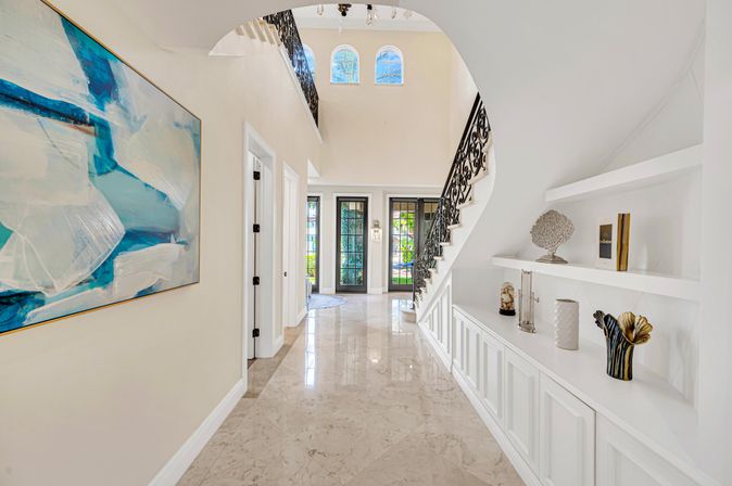 Bright luxury foyer with polished marble floor, curved staircase with ornate wrought-iron railing, built-in white display shelves with decorative vases, large abstract blue painting and glass double front doors letting in natural light.