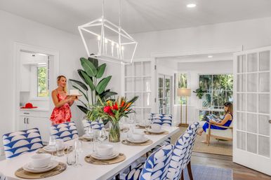 Sunlit open-plan dining room with a long white table set for eight, blue patterned chairs, tropical floral centerpiece and pendant light; a woman in a red sundress brings a cake from the kitchen while another relaxes reading in the adjacent sunroom through glass French doors.