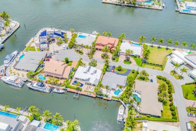 Aerial view of a sun-soaked coastal canal neighborhood with waterfront homes, palm trees, private docks, yachts, and backyard pools.