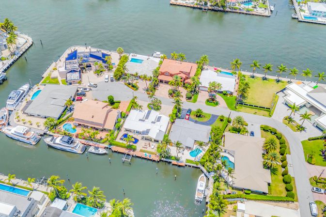Aerial view of a sun-soaked coastal canal neighborhood with waterfront homes, palm trees, private docks, yachts, and backyard pools.