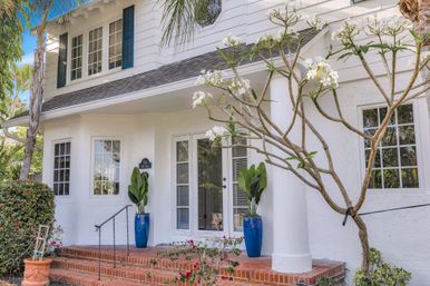 Sunny coastal-style white cottage front porch with brick steps, blue ceramic planters, tropical foliage and a blooming plumeria tree.
