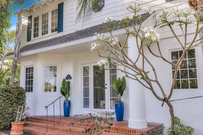 Sunny coastal-style white cottage front porch with brick steps, blue ceramic planters, tropical foliage and a blooming plumeria tree.