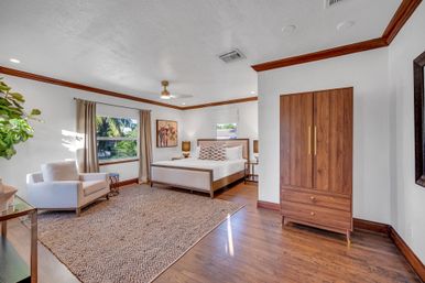Sunlit spacious master bedroom with king bed, neutral linens and patterned pillows, wooden floors, walnut armoire, upholstered armchair, textured area rug and large window framing palm trees