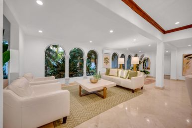 Coastal-chic living room with arched windows framing a lush tropical garden, white sofas and armchairs, patterned area rug, low coffee table, marble floors and recessed lighting.