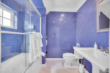 Vibrant blue mosaic-tiled bathroom with glass-enclosed tub-shower, white pedestal sink, toilet, and folded towels.
