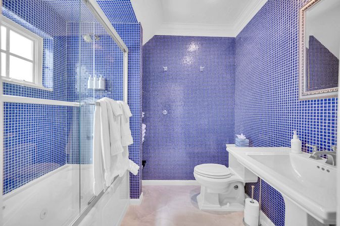 Vibrant blue mosaic-tiled bathroom with glass-enclosed tub-shower, white pedestal sink, toilet, and folded towels.