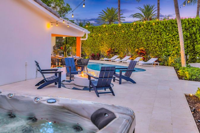 Twilight tropical backyard patio with hot tub in foreground, circular fire pit surrounded by Adirondack chairs, pool and lounge chairs, string lights and palm trees.