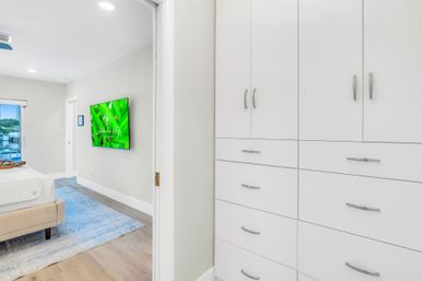 Bright modern coastal bedroom seen from a hallway with white built-in cabinets and drawers, an upholstered bed on a blue area rug, wall-mounted TV showing green screen, hardwood floors, and a waterfront view through the window.
