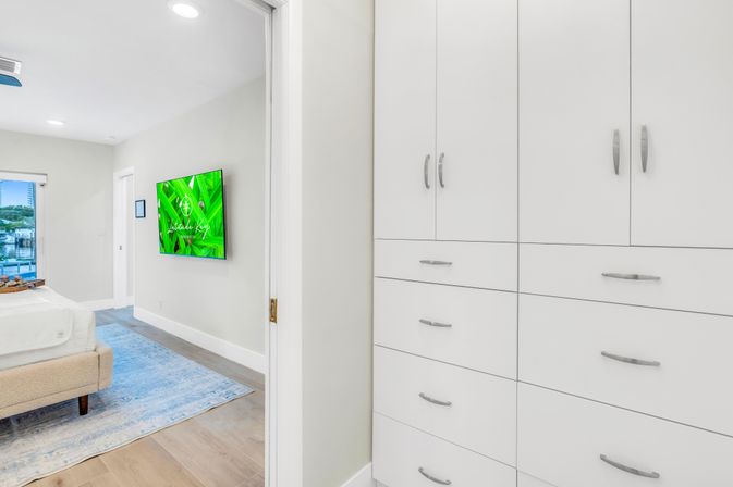 Bright modern coastal bedroom seen from a hallway with white built-in cabinets and drawers, an upholstered bed on a blue area rug, wall-mounted TV showing green screen, hardwood floors, and a waterfront view through the window.