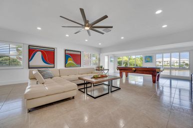 Bright waterfront game room with beige leather sectional, modern coffee table and ceiling fan, pool table by floor-to-ceiling windows overlooking palm-lined canal and marina, colorful abstract art