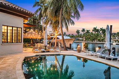 Luxury tropical waterfront backyard at sunset with a curved pool reflecting string lights, swaying palm trees, lounge chairs, a thatched tiki hut and a docked yacht on a calm canal