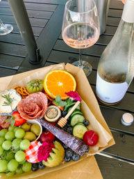 Colorful charcuterie box with salami rose, cheese, olives, grapes, kiwi, orange slice, cucumber and honey paired with a chilled rosé glass and bottle on a dark outdoor patio table