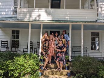 Smiling group of eight friends holding wine glasses on the steps of a white wooden farmhouse porch with rocking chairs, potted plants and green shrubs in bright summer light.
