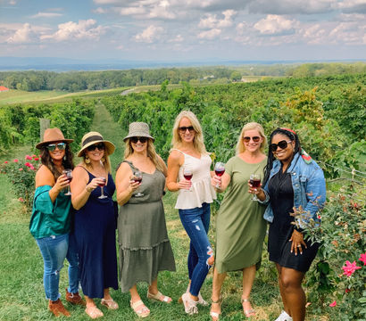 Six women raise glasses of red wine in a sunny vineyard, lush grapevines stretching into rolling green countryside under a dramatic blue sky with puffy clouds.