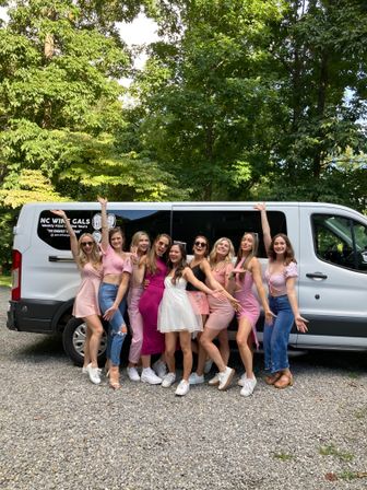 Group of friends in pink and white outfits posing happily in front of a white shuttle van on a gravel drive with leafy trees in the background, upbeat bachelorette party vibe.