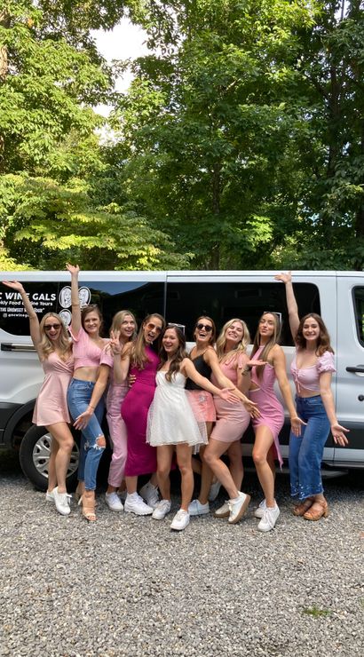 Group of friends in pink and white outfits posing happily in front of a white shuttle van on a gravel drive with leafy trees in the background, upbeat bachelorette party vibe.