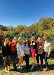 Group of seven women in an outdoor rose garden on a sunny autumn day, wearing heart-shaped sunglasses amid red roses, vineyard-style rows and colorful fall trees under a clear blue sky.