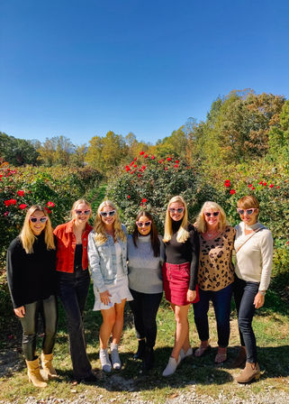 Group of seven women in an outdoor rose garden on a sunny autumn day, wearing heart-shaped sunglasses amid red roses, vineyard-style rows and colorful fall trees under a clear blue sky.