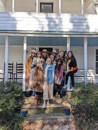 Smiling women in fall coats and hats pose together on the steps of a white farmhouse-style front porch.