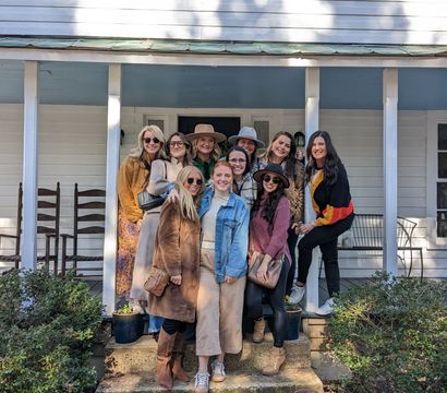 Smiling women in fall coats and hats pose together on the steps of a white farmhouse-style front porch.