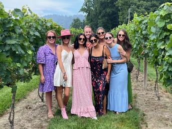 Eight women in colorful summer dresses and sunglasses posing and smiling between rows of green grapevines in a sunlit vineyard with rolling hills in the background.