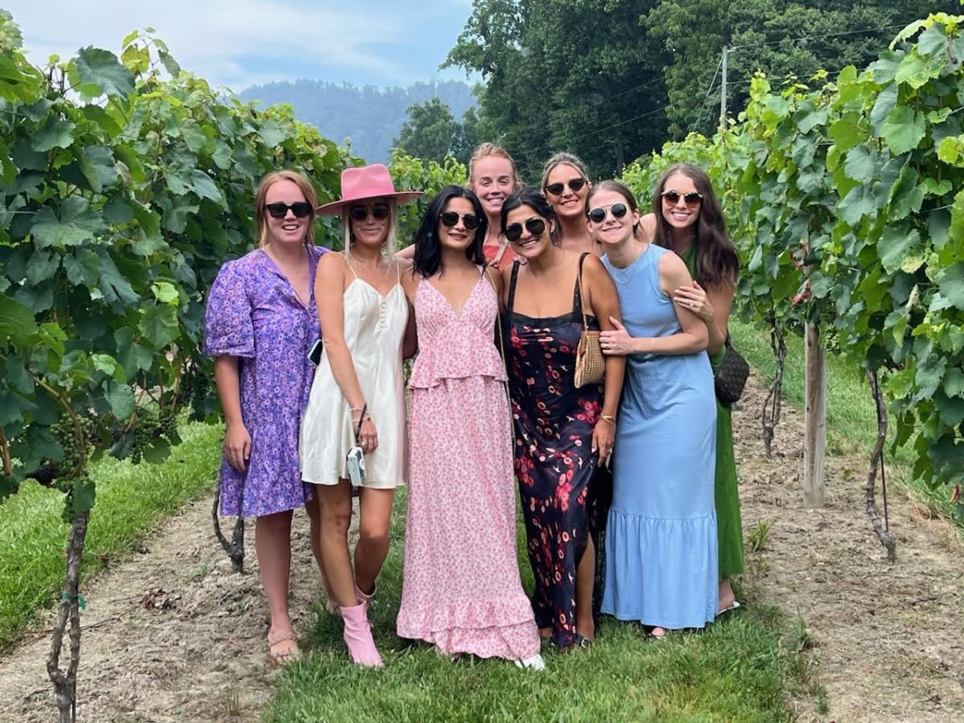 Eight friends in colorful summer dresses and sunglasses posing between vineyard rows, lush grapevines and tree‑covered hills in the background — sunny winery outing.
