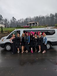 Nine women in black dresses with colorful fascinators smiling and posing in front of a white wine-tour van beside a grassy hillside and pavilion on an overcast day