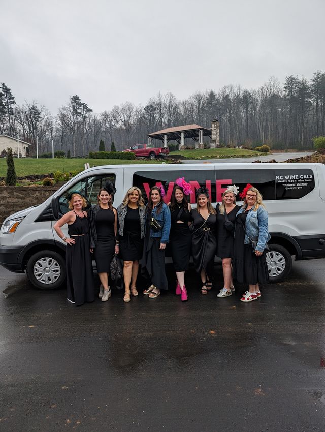 Nine women in black dresses and playful shoes posing and smiling in front of a white wine-tour van on a rainy day at a wooded hilltop winery estate
