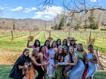 About 14 women smiling and holding wine glasses at a vineyard wine tasting, standing in front of neat vine rows, rolling hills and a bright blue sky; one holds a small dog.