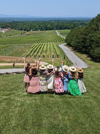 Group of women in sundresses and straw hats with arms raised, overlooking neat rows of a vineyard, winding country road and rolling hills on a sunny day