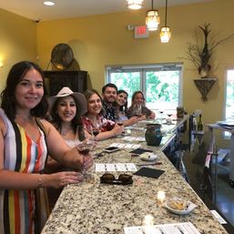 Smiling group enjoying a wine tasting at a bright winery tasting room bar, holding red wine glasses over a speckled granite countertop with tasting notes and small snacks