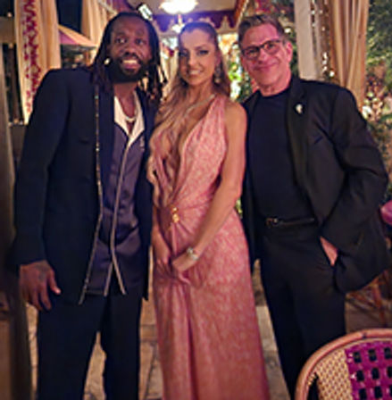 Three well-dressed adults — two men and a woman in a pink evening gown — smiling at an elegant outdoor patio evening party with draped curtains and string lights.