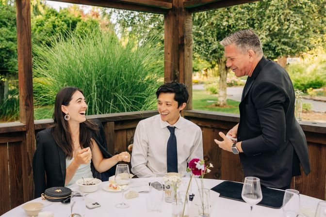Smiling diners at an outdoor garden patio: a woman in a black jacket and a man in a white shirt with a tie sit at a table with wine glasses and dessert while a server in a dark suit entertains them under a wooden gazebo.