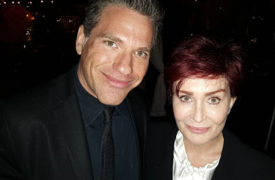 Close-up selfie of two smiling adults — a man with salt-and-pepper hair and a woman with short auburn hair — wearing black formal attire at a dimly lit evening event.