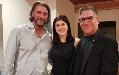 Casual group photo of three adults smiling indoors in a cozy home setting — bearded man in a light button-up, woman in a black sleeveless top, and a man with glasses in a dark blazer under a wooden ceiling beam.