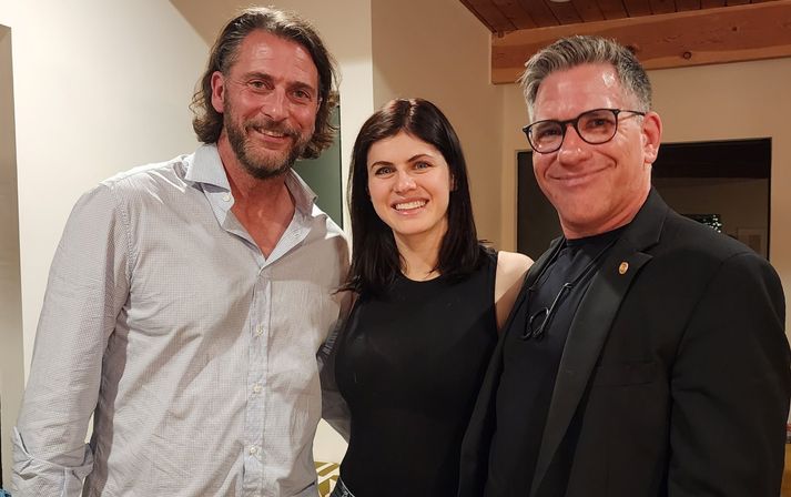Casual group photo of three adults smiling indoors in a cozy home setting — bearded man in a light button-up, woman in a black sleeveless top, and a man with glasses in a dark blazer under a wooden ceiling beam.