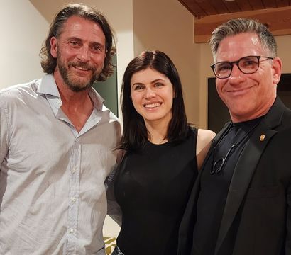 Casual group photo of three adults smiling indoors in a cozy home setting — bearded man in a light button-up, woman in a black sleeveless top, and a man with glasses in a dark blazer under a wooden ceiling beam.
