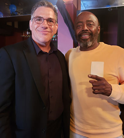 Two smiling men posing in a dimly lit bar or lounge — one in glasses and a dark suit, the other in a tan sweatshirt pointing at a small white card, casual nightlife scene.