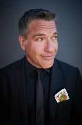 Close-up portrait headshot of a man in a black suit and tie with a Jack of Hearts playing card tucked in his pocket, wearing a wry smile and looking off to the side against a neutral gray background.