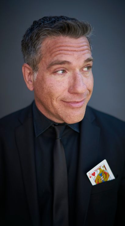 Close-up portrait headshot of a man in a black suit and tie with a Jack of Hearts playing card tucked in his pocket, wearing a wry smile and looking off to the side against a neutral gray background.