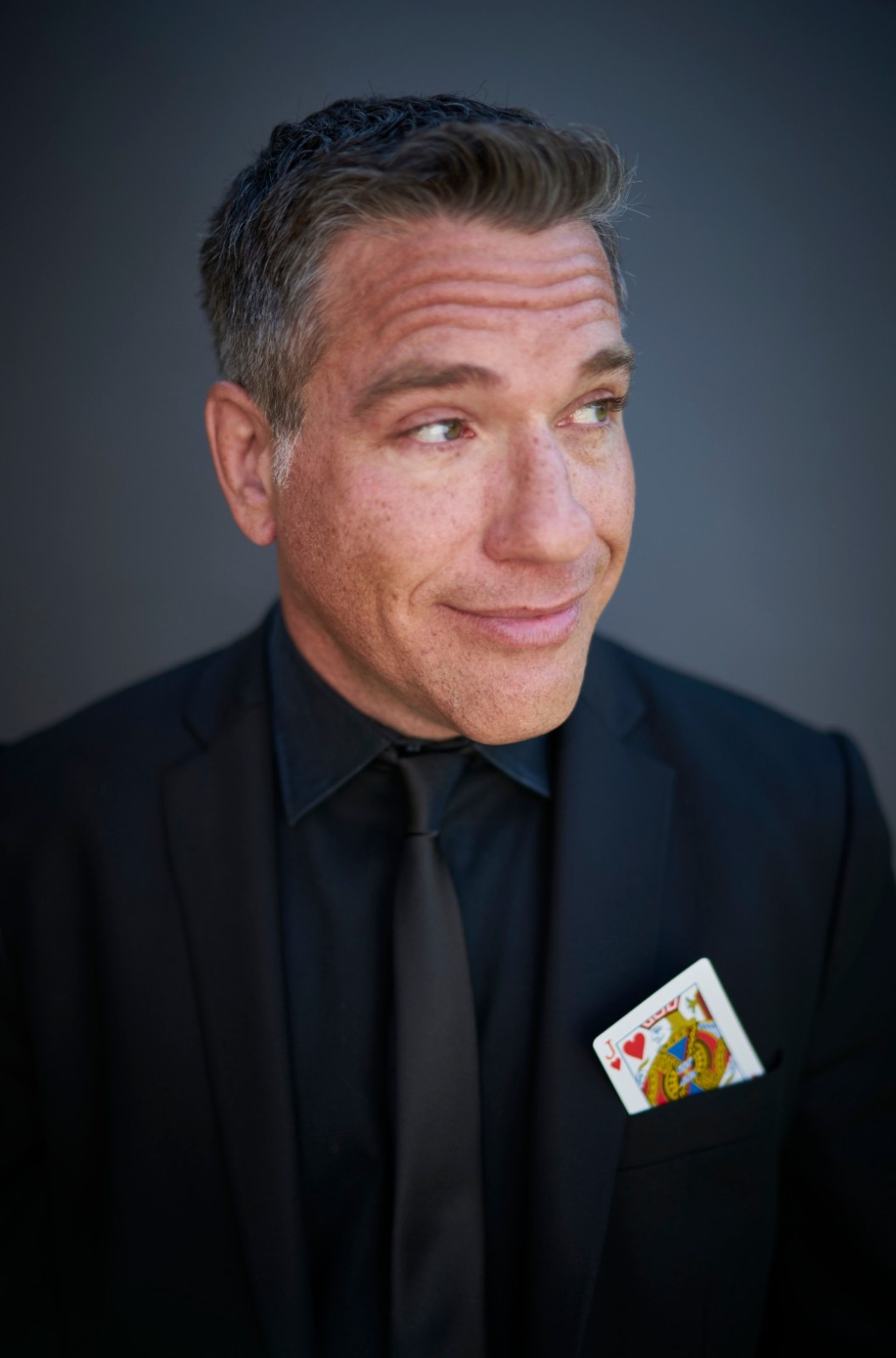 Close-up portrait headshot of a man in a black suit and tie with a Jack of Hearts playing card tucked in his pocket, wearing a wry smile and looking off to the side against a neutral gray background.