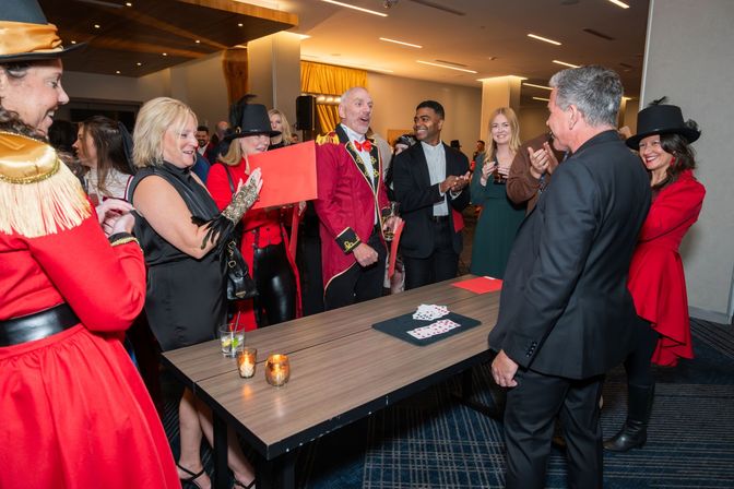 Crowd gathered around a table in a hotel event space as a ringmaster-style entertainer in a red coat performs a card trick, guests in red costumes and formalwear cheer with drinks and candles on the table.