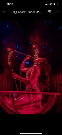 Cabaret performer kneeling in a giant glass bowl on a nightclub stage, fire-eating with flaming torches under moody red lighting — burlesque-style dinner show vibe.