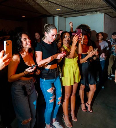 Group of people at a lively indoor club or concert, standing near the stage, smiling and recording the performance on smartphones under colorful lights.