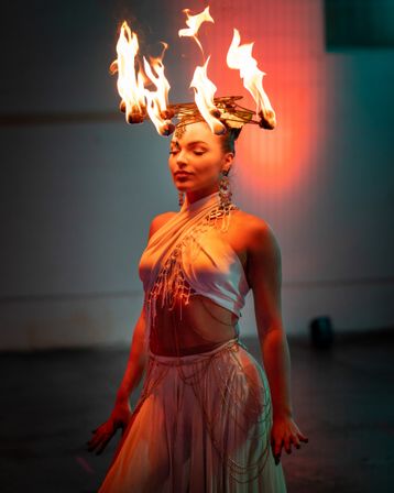 Fire dancer in flowing white costume wearing a crown of flames, eyes closed amid dramatic indoor stage lighting