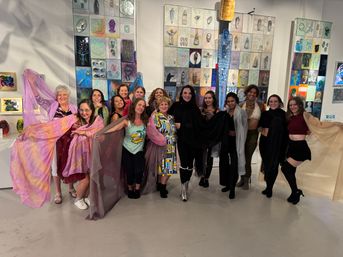 Group of women posing at a contemporary art gallery event in front of colorful mixed-media wall panels, holding flowing sheer scarves and smiling.