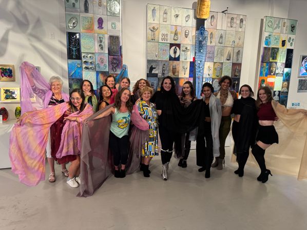 Group of women posing at a contemporary art gallery event in front of colorful mixed-media wall panels, holding flowing sheer scarves and smiling.
