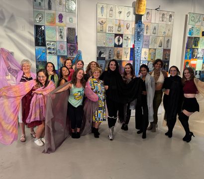Group of women posing at a contemporary art gallery event in front of colorful mixed-media wall panels, holding flowing sheer scarves and smiling.