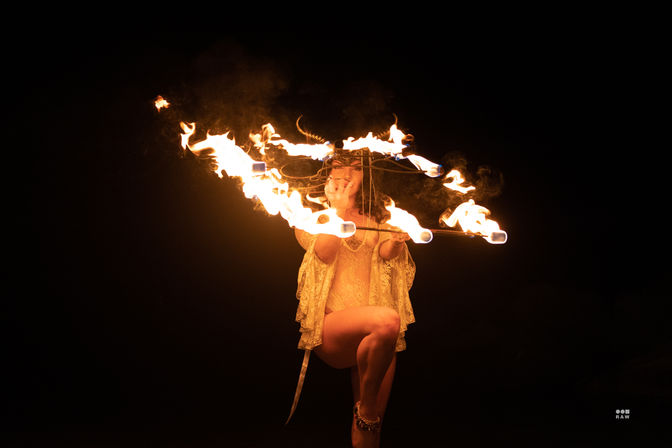 Fire performer balancing on one leg while twirling a flaming hoop and torches in a dramatic nighttime show