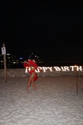 Dancer in a red costume performing on a sandy beach at night in front of illuminated HAPPY BIRTHDAY letters, with torches and distant city lights in the background.